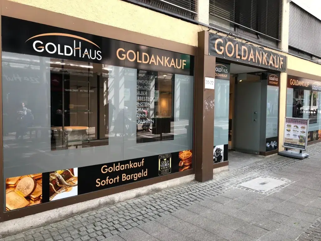 Geschäftsfassade mit goldener Aufschrift "Goldankauf", große Fenster und Werbeplakate über Schmuck.