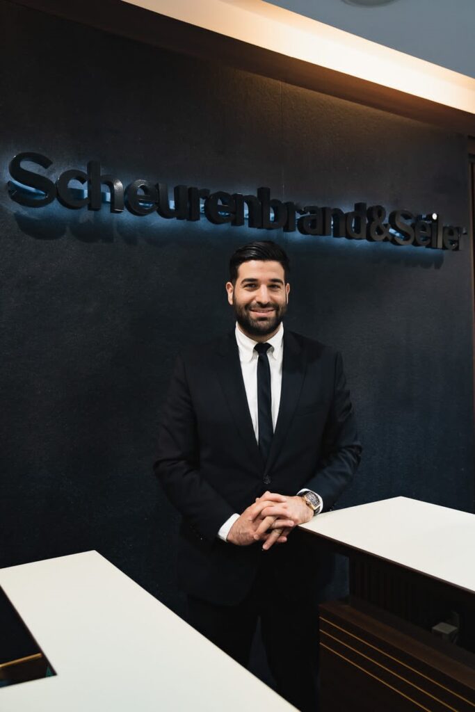 Ein lächelnder Mann in Anzug vor dunkler Wand mit Firmenlogo.